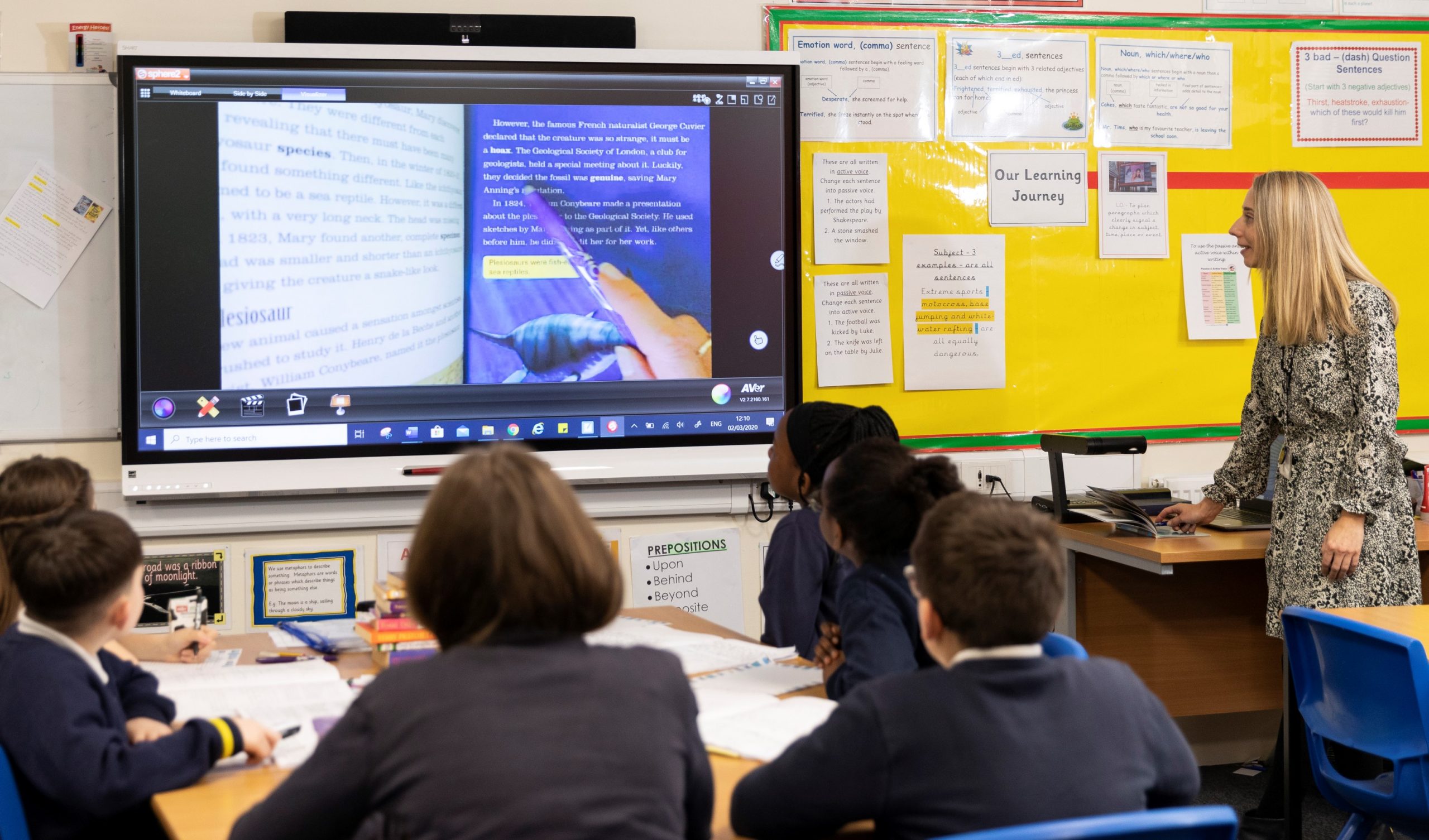 A teacher uses a visualiser to enlarge a book onto the interactive whiteboard so pupils can read with her.
